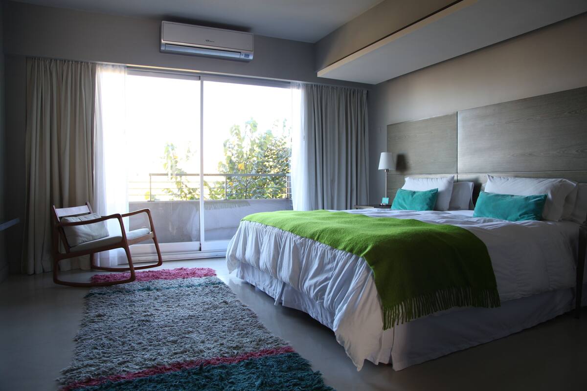 Un dormitorio moderno con una cama grande, un sillón mecedor, un balcón con cortinas y vistas a zonas verdes.