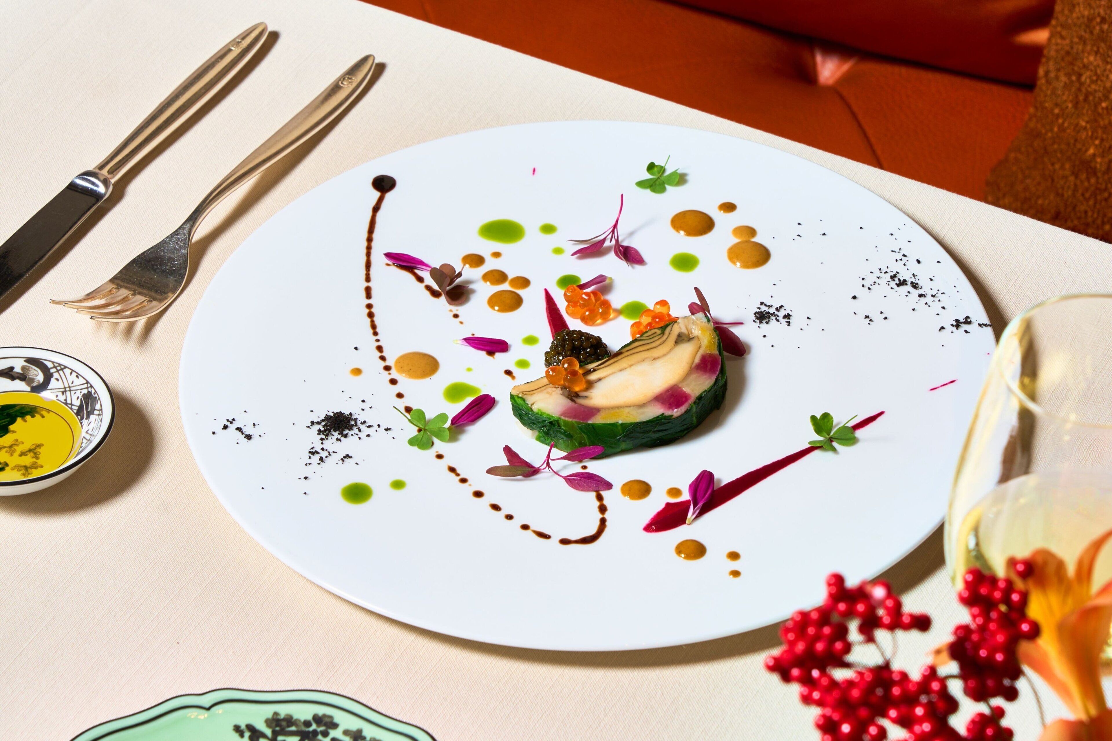A white plate with a colorful dessert, a fork, a knife, and a small bowl of sauce on a table.