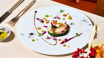 A white plate with a colorful dessert, a fork, a knife, and a small bowl of sauce on a table.