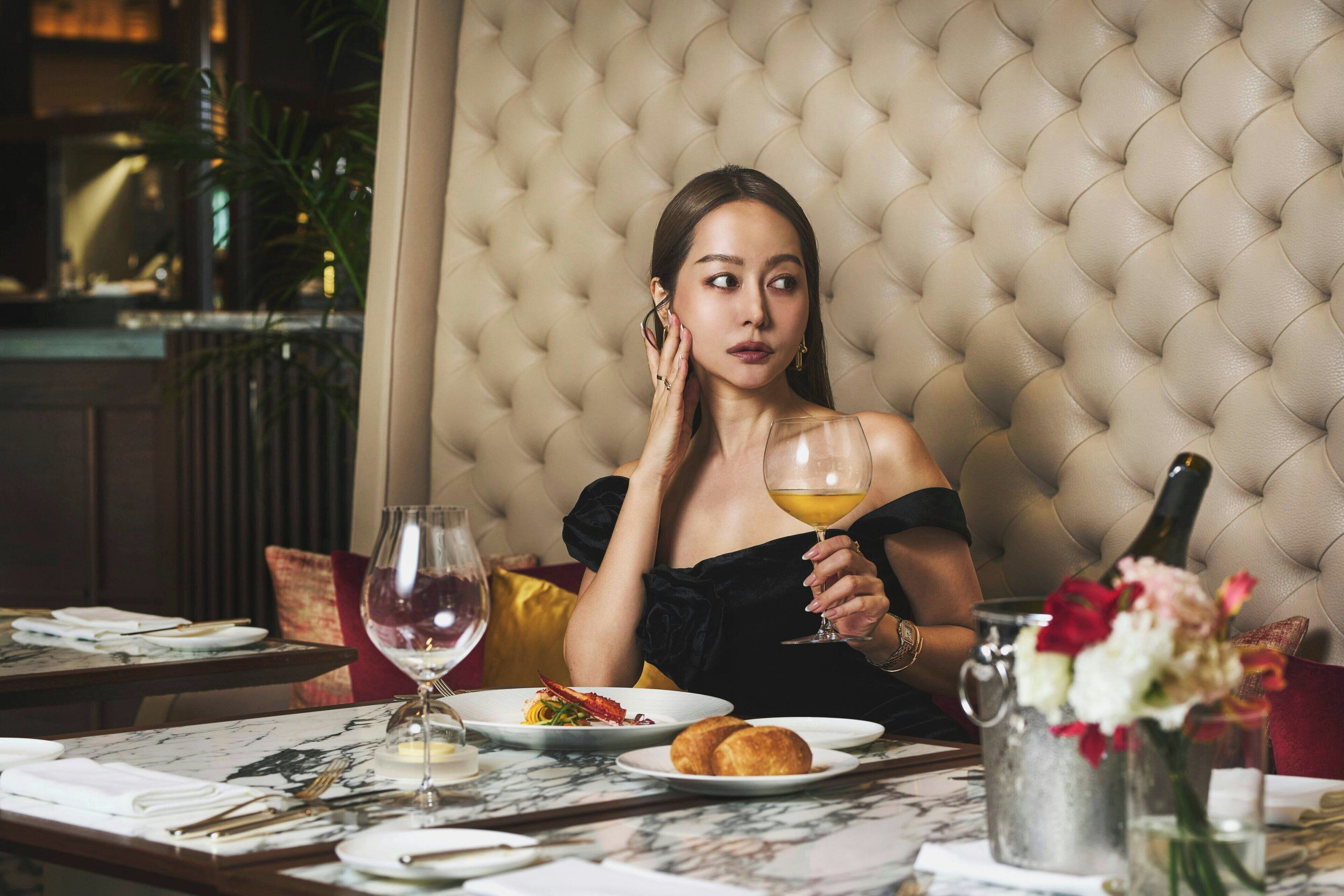 Une femme est assise à une table devant une bouteille de champagne, des verres à vin et un vase de fleurs.