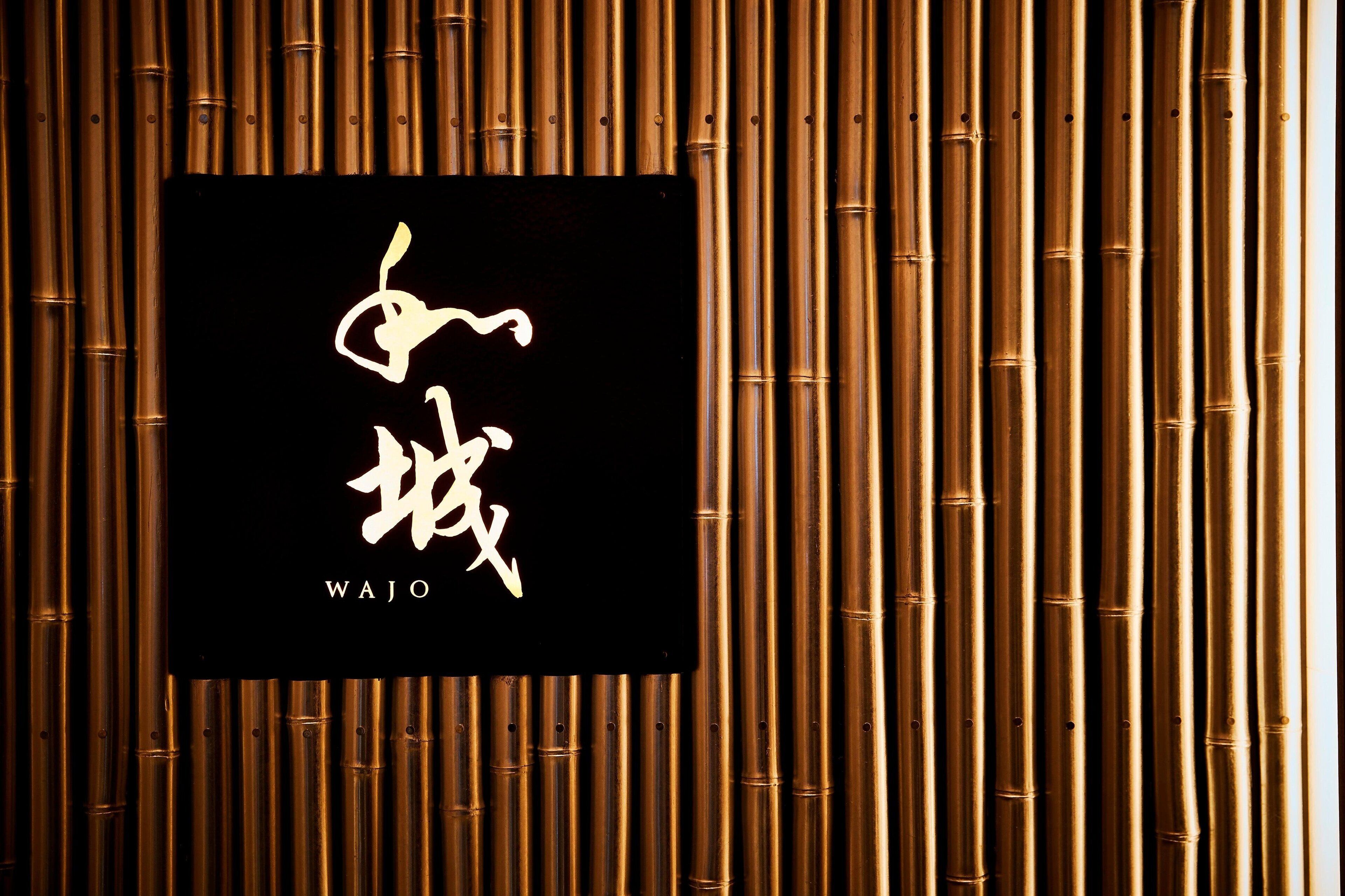 A black sign with gold Chinese characters and the word "WAJO" on a bamboo background.