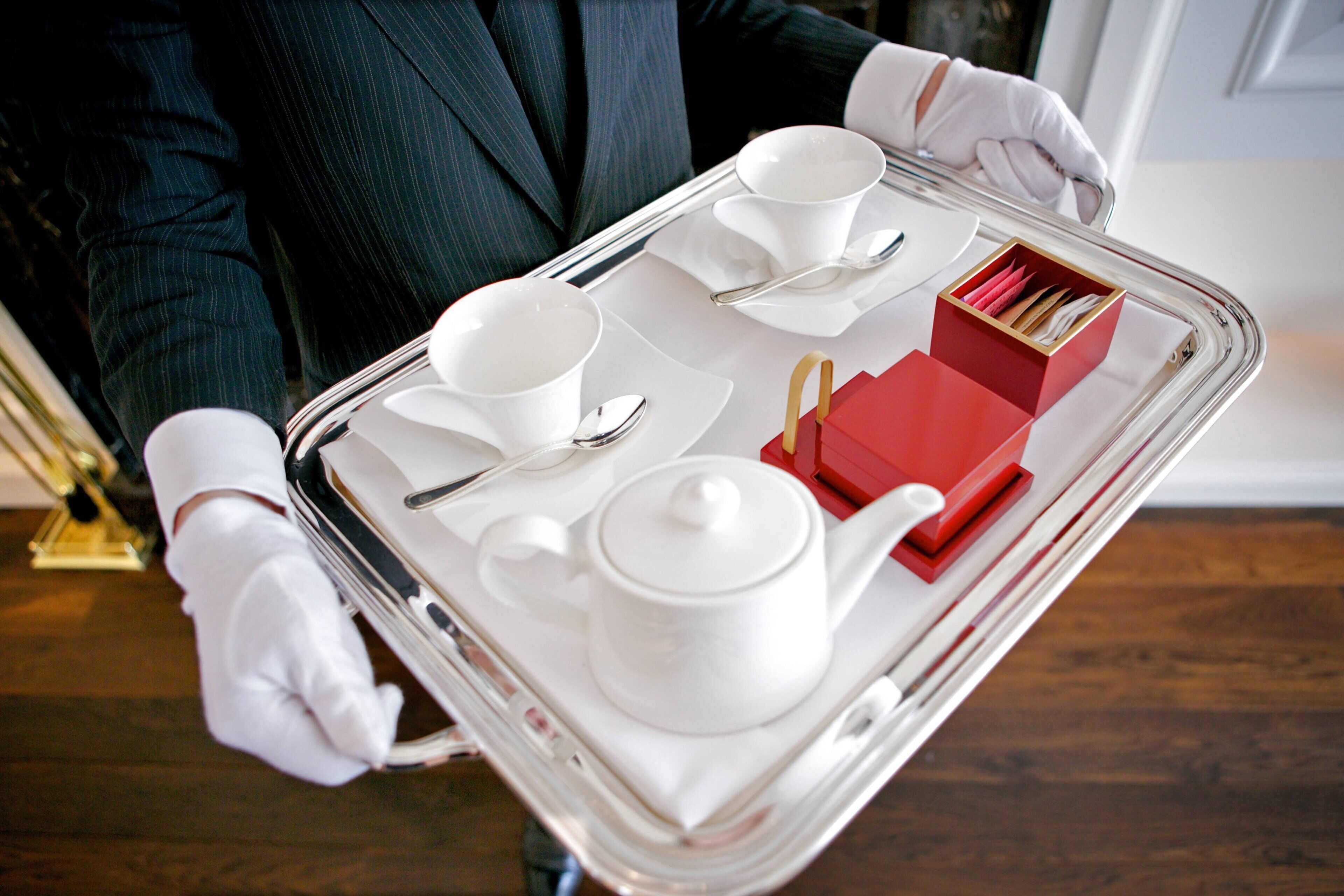 A person in a dark suit and white gloves holding a tray with white teacups, a teapot, and a red box of tissues.
