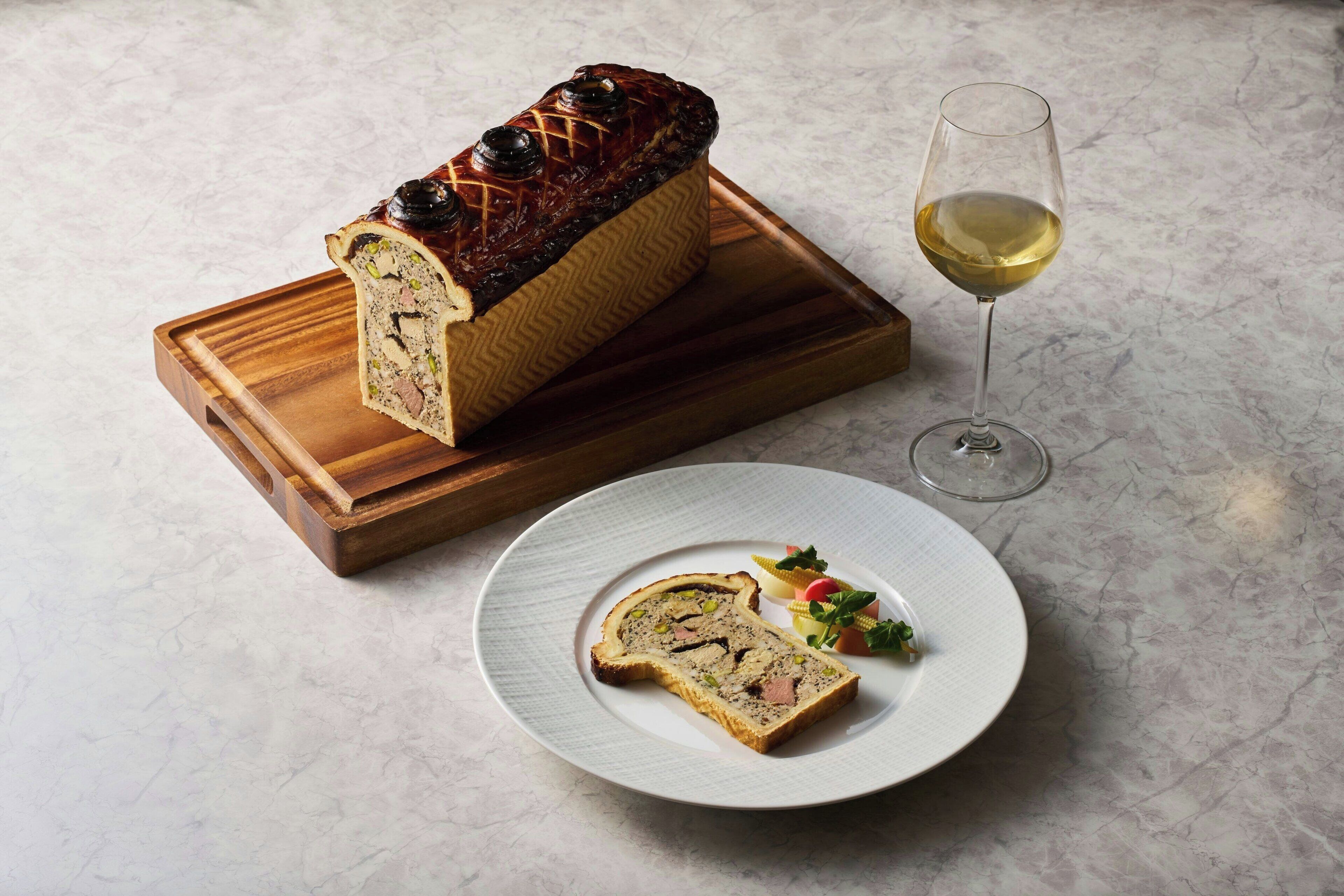 A loaf of bread on a wooden board, a glass of white wine, and a plate with bread slices and vegetables.
