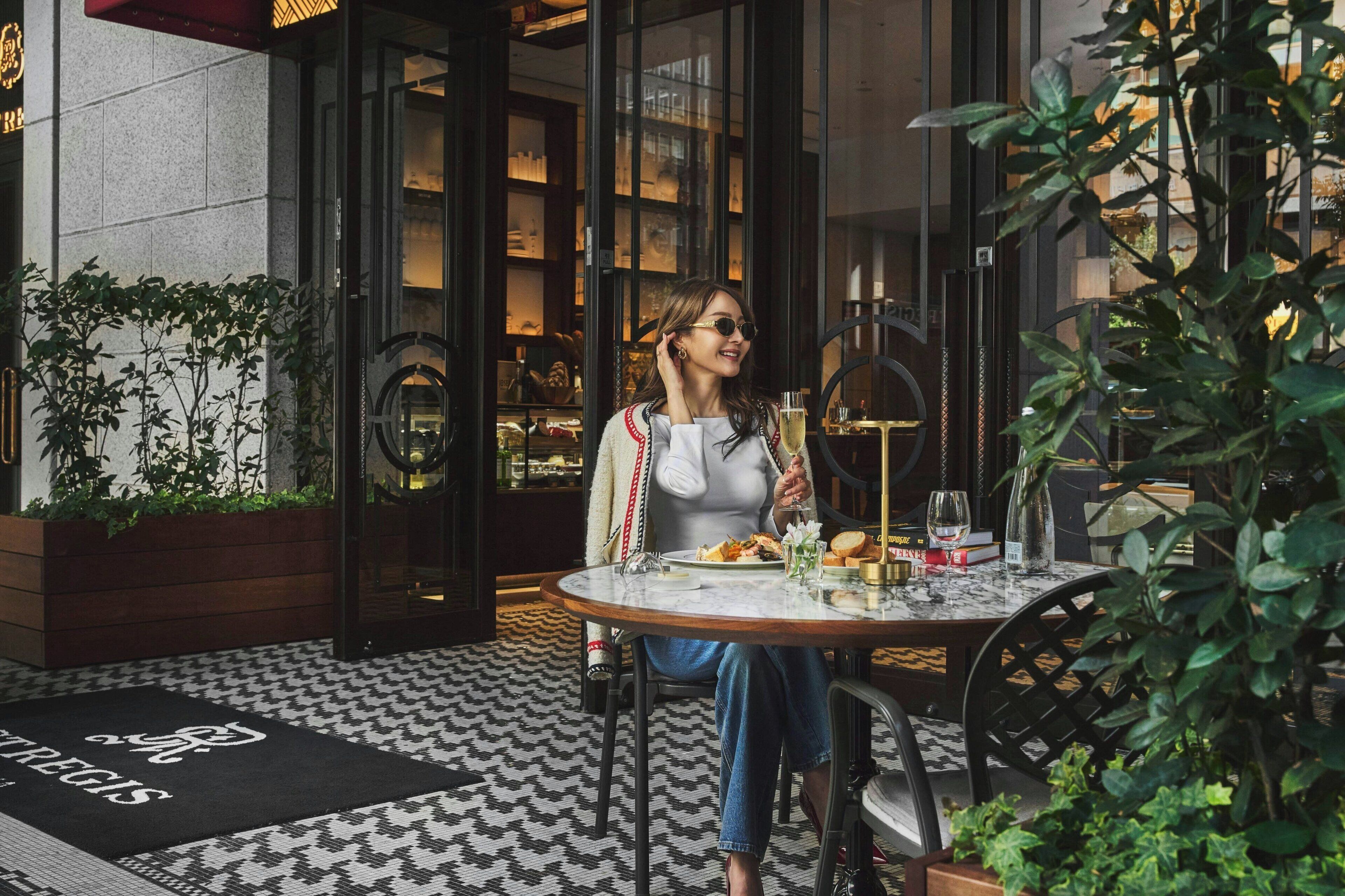 Una mujer cenando en una mesa al aire libre con una bebida en mano, rodeada de follaje y un suelo con diseño.