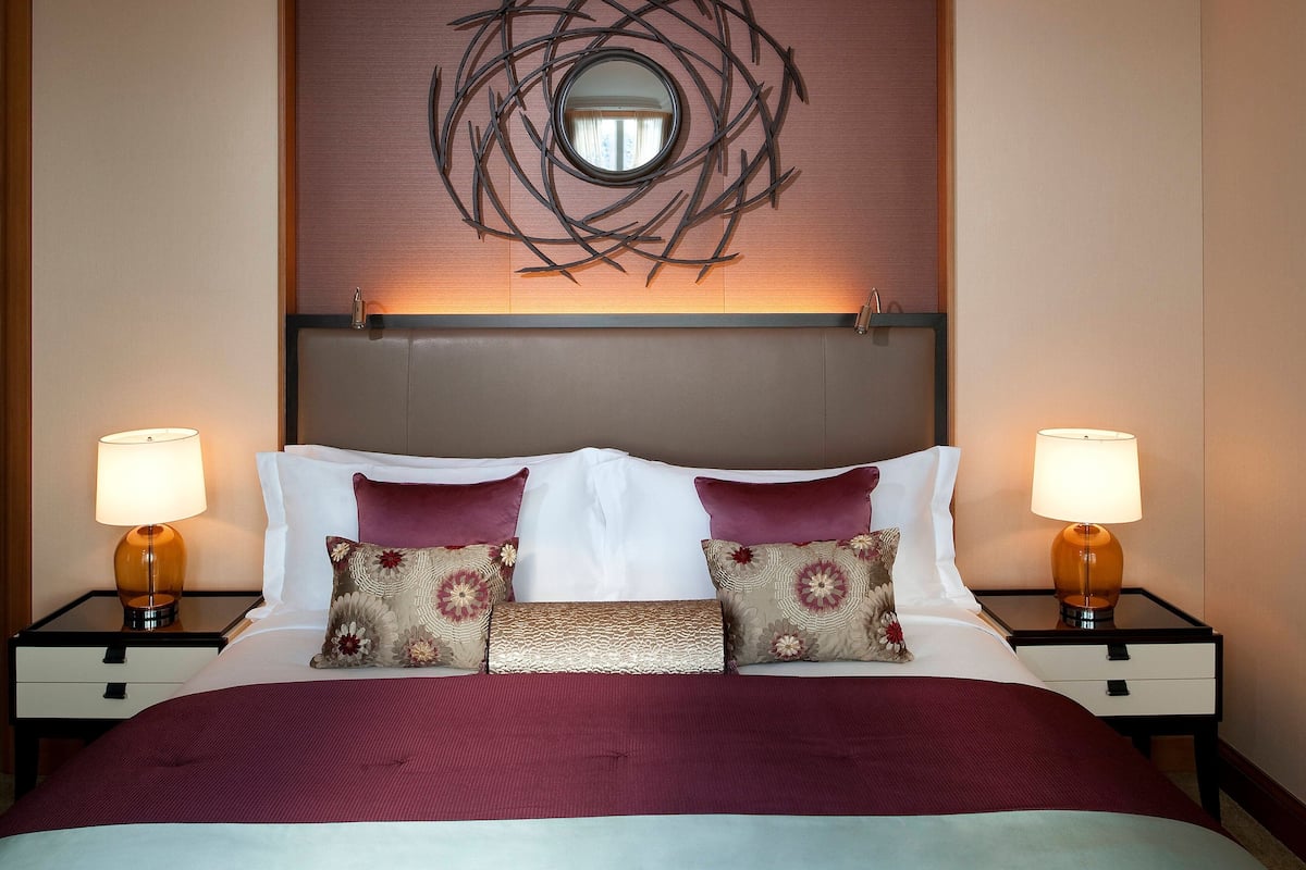 A neatly made bed with a red bedspread, two decorative pillows, and a round mirror above the headboard. There are two bedside lamps on nightstands.