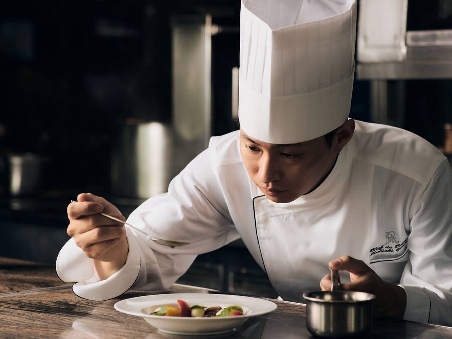 Un chef vêtu d’une blouse et d’un chapeau blancs, tenant une cuillère et une casserole, avec une assiette de nourriture devant lui.