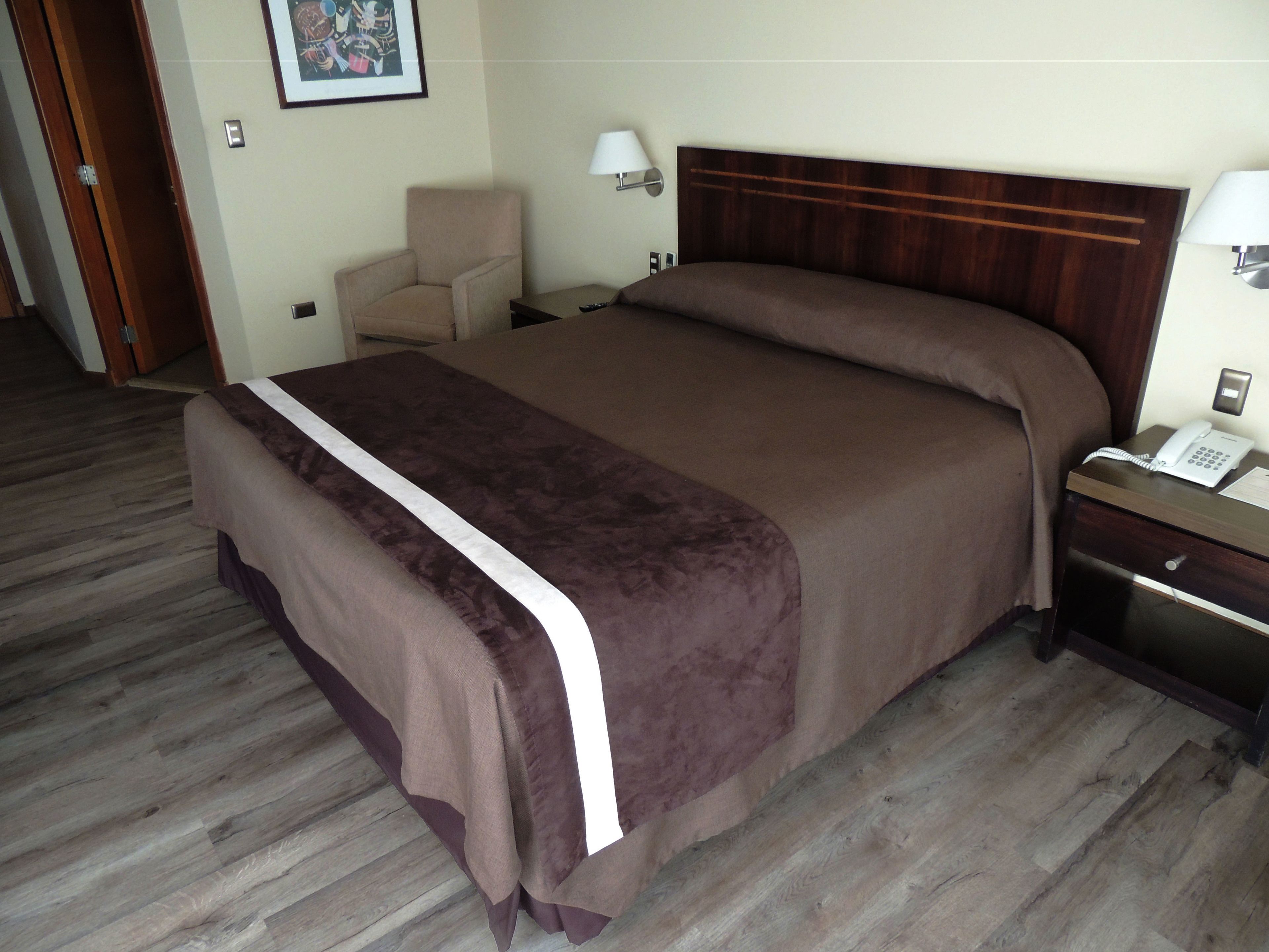 A hotel room with a bed, a chair, a nightstand with a phone, and a lamp.