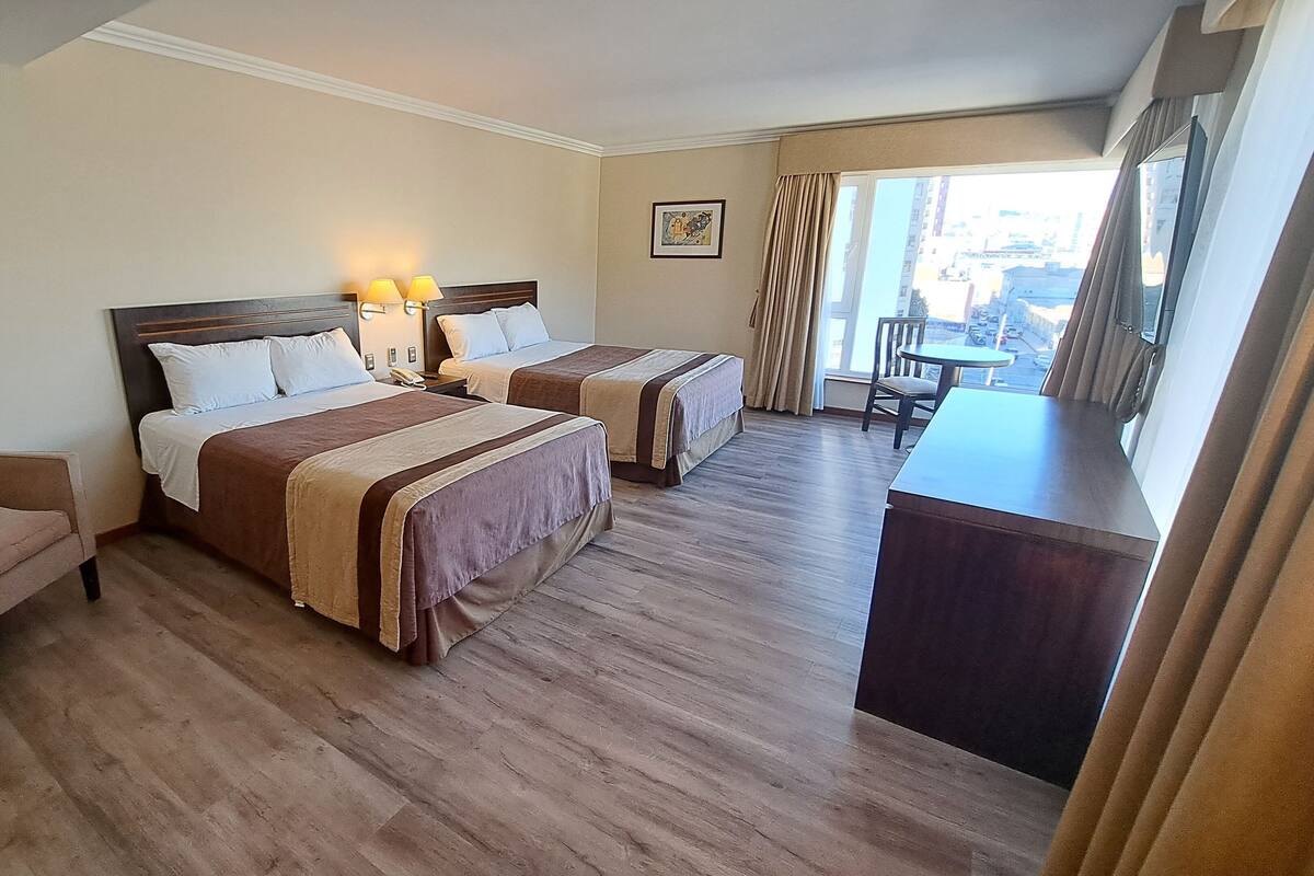 A hotel room with two beds, a desk, a chair, and a large window with curtains.