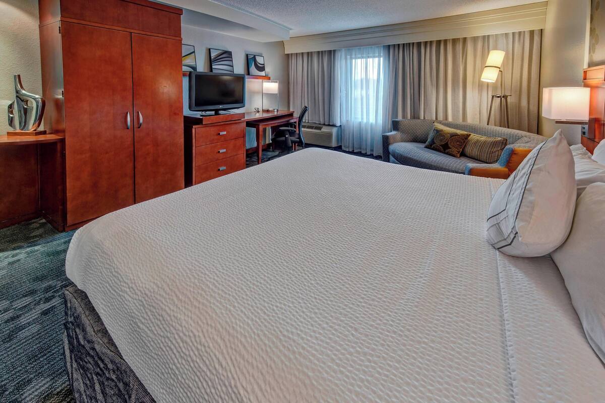 A hotel room with a large bed, a desk with a television, a chair, a sofa, and a lamp.