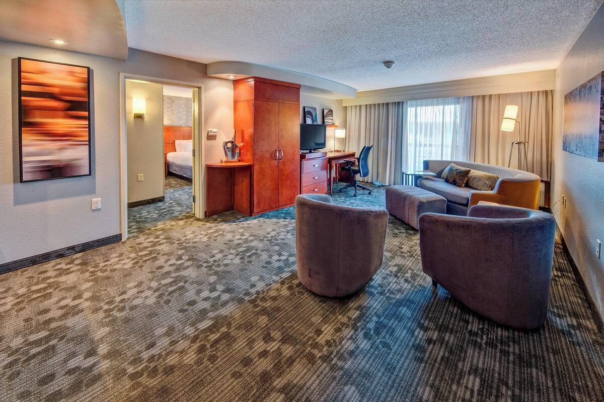 A hotel room with a large bed, a desk with a computer, a TV, a sofa, and a carpeted floor.