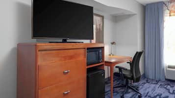 A hotel room with a flat-screen TV on a wooden dresser, a microwave, a small refrigerator, a desk with a chair, and a window with curtains.