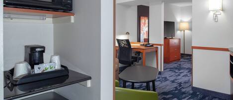 A hotel room with a microwave, coffee maker, and a small refrigerator on the left. The room has a desk, chair, and a lamp.