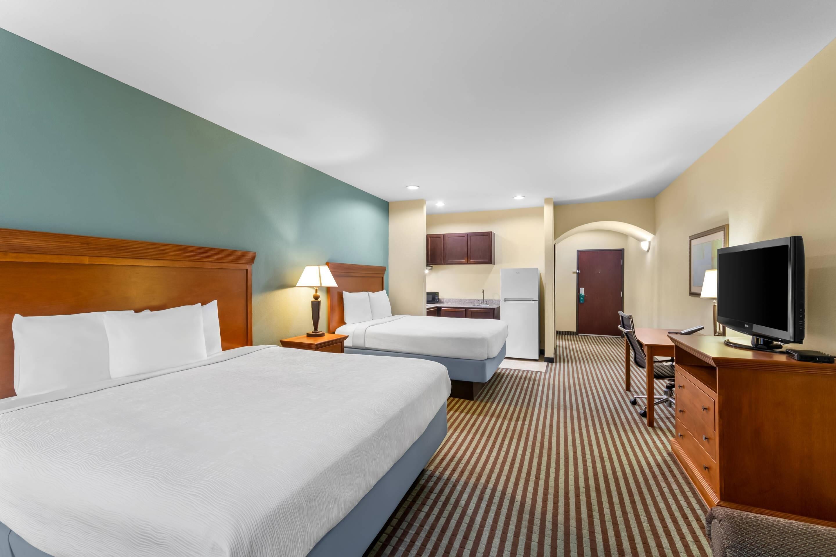 A hotel room with two beds, a desk, a TV, and a kitchenette.