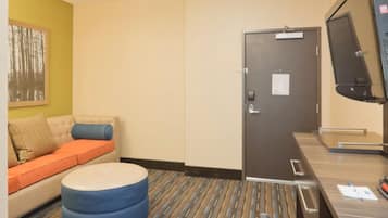 A hotel room with a sofa, ottoman, desk, and a door with a monitor.
