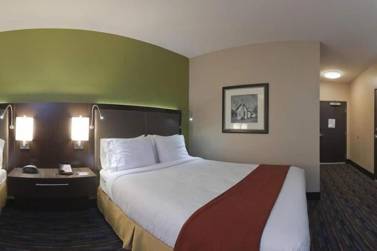 A hotel room with two beds, a headboard, a bedside table with a lamp, a framed picture on the wall, and a carpeted floor.