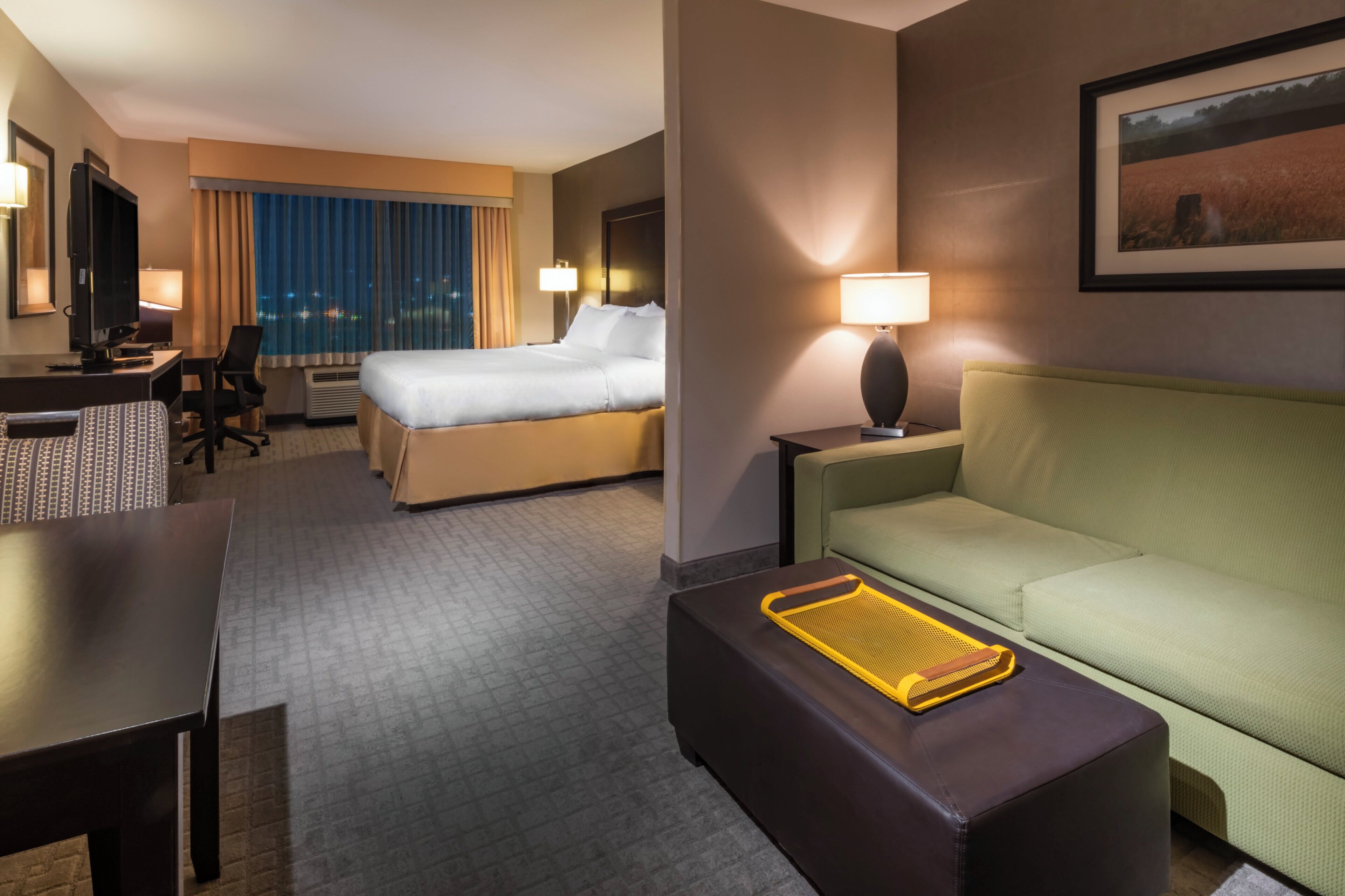 A hotel room with a bed, a desk, a chair, a sofa, a lamp, and a painting on the wall.