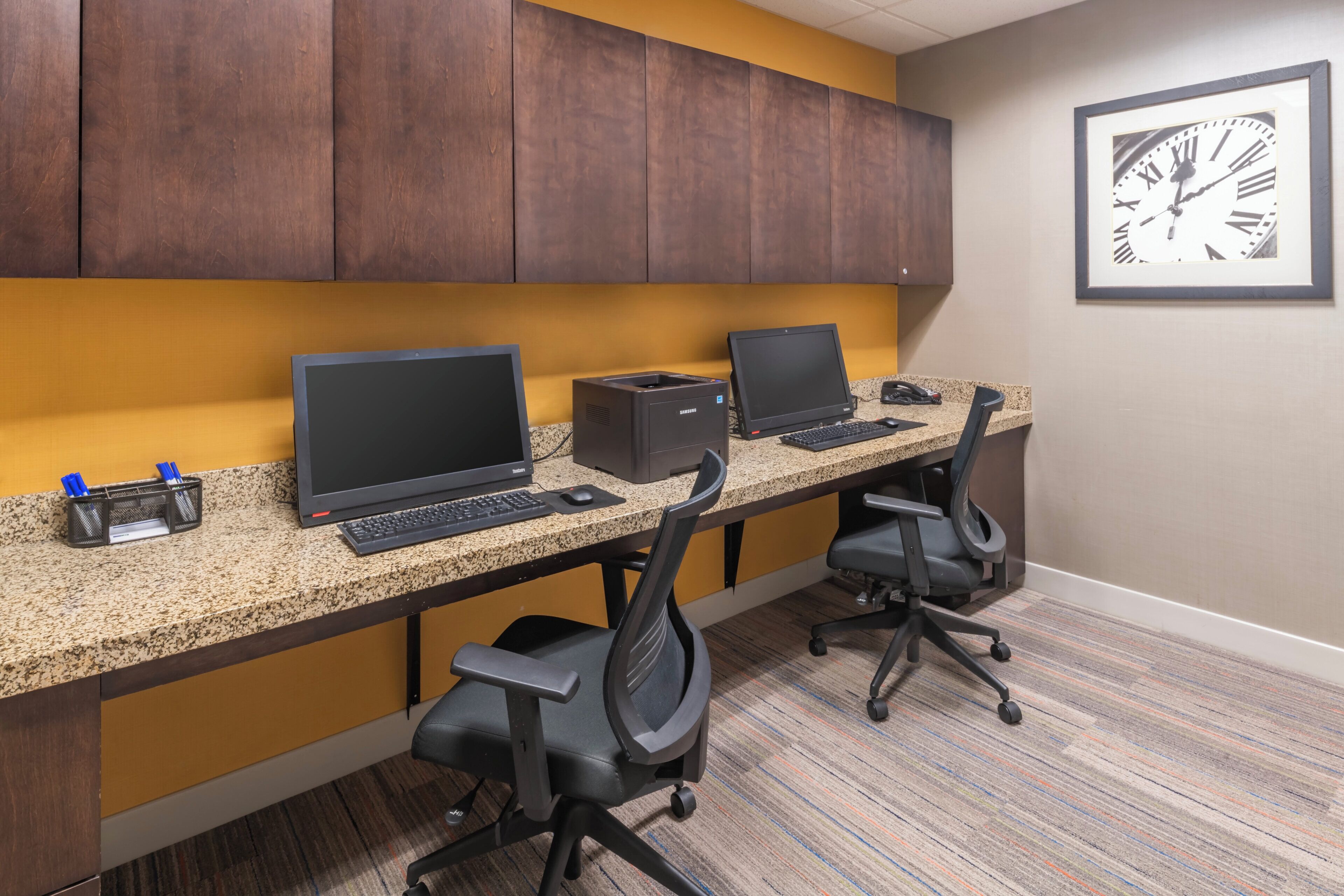 A workspace with two desks, each with a laptop, keyboard, and mouse. There are office chairs and a framed clock on the wall.
