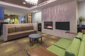 A hotel lobby with a sofa, a coffee table, a flat-screen TV, a fireplace, and a patterned carpet. - Holiday Inn Express Hotel & Suites Hays by IHG (Hays)