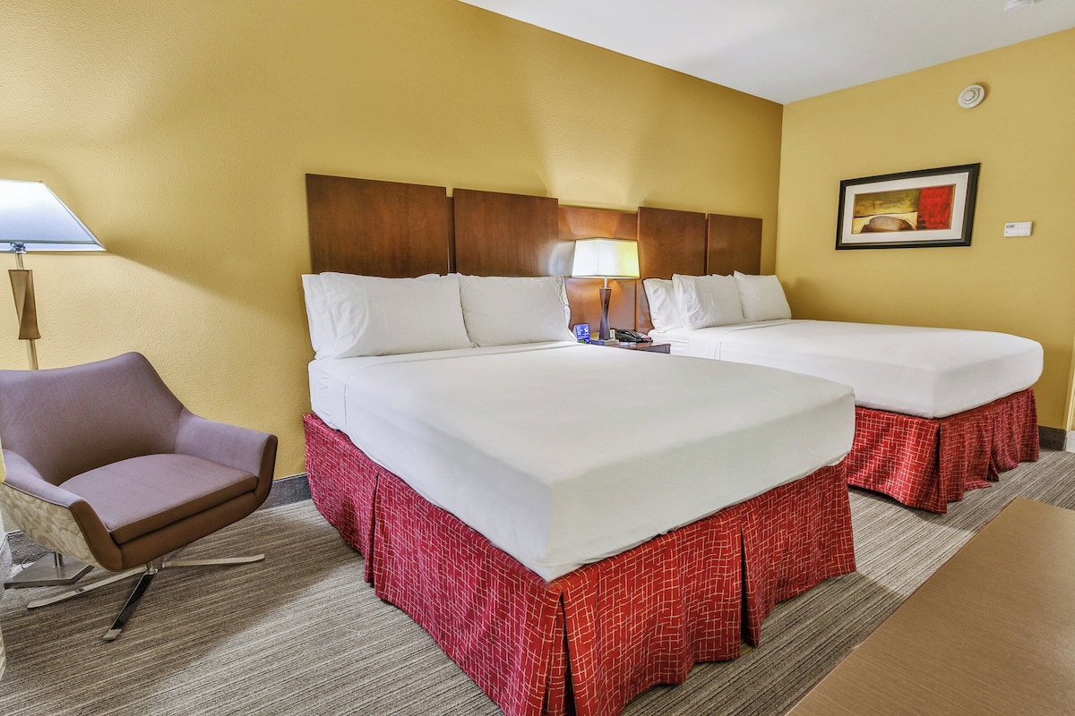 A hotel room with two beds, a chair, a lamp, and a painting on the wall.