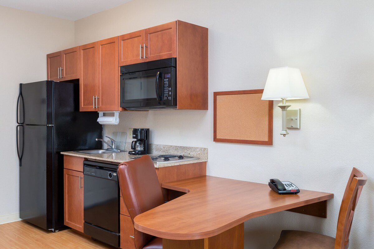 A compact kitchen with wooden cabinets, a black refrigerator, a microwave, a coffee maker, and a table with a chair.