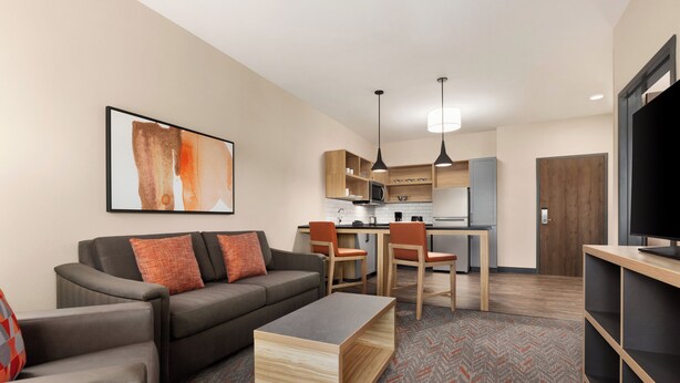 A modern living room with a sofa, coffee table, and TV stand. There is a kitchen area with cabinets and a microwave.
