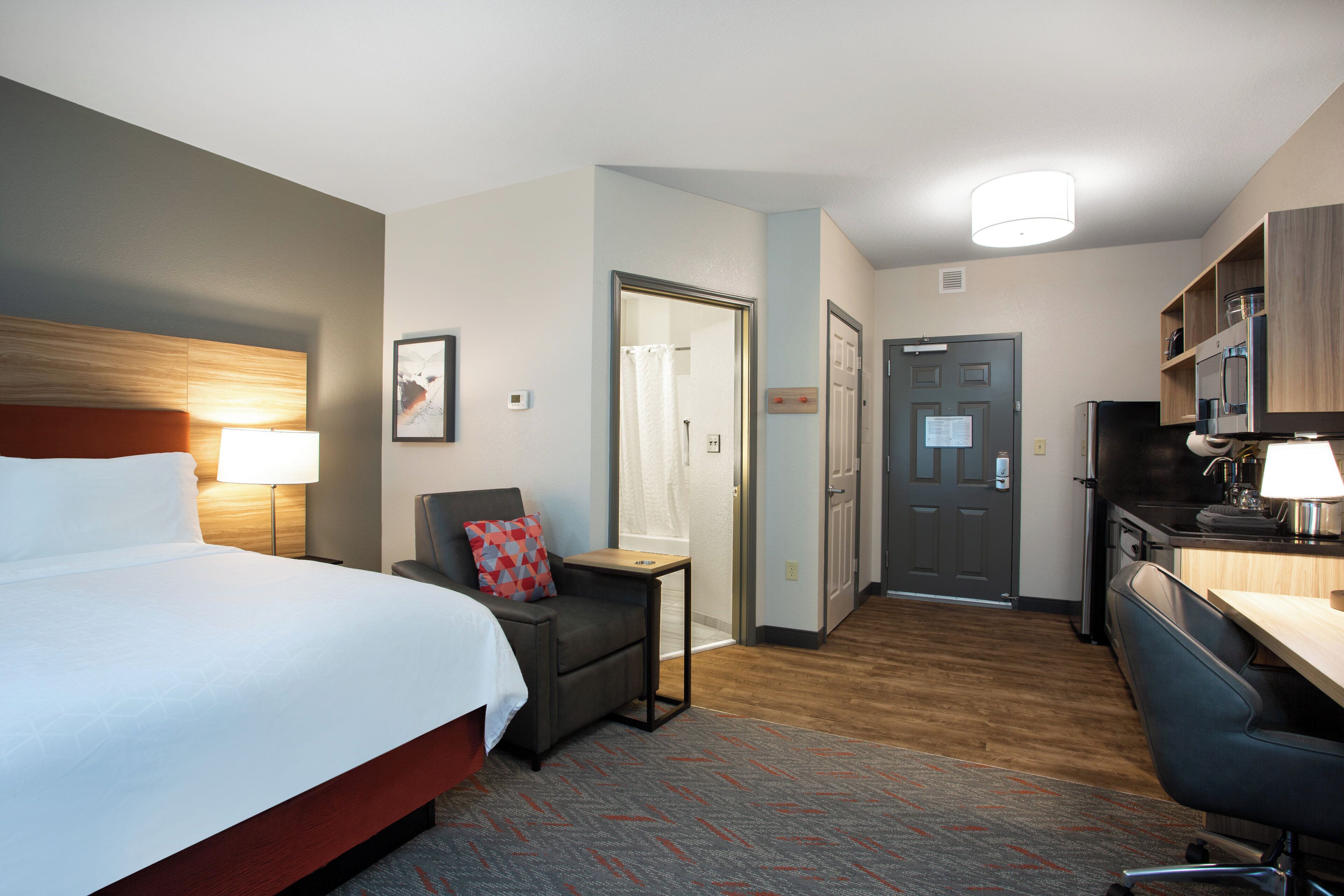 A hotel room with a bed, a desk, a chair, a small table, a shower, and a kitchenette.