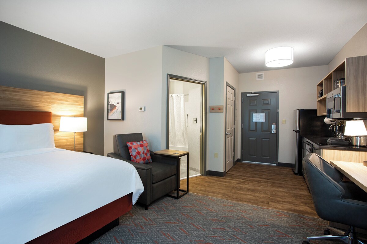 A hotel room with a bed, a desk, a chair, a small table, a shower, and a kitchenette.