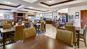 Un grand espace de salle à manger avec plusieurs tables et chaises, un coin cuisine avec un panneau indiquant « DÉCHETS », et un téléviseur fixé au mur.