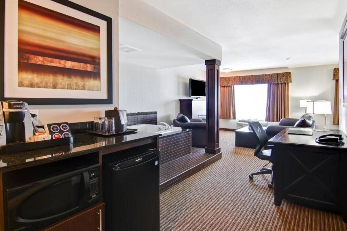 A hotel room with a kitchenette, a flat-screen TV, a sofa, a desk, and a large window with curtains.
