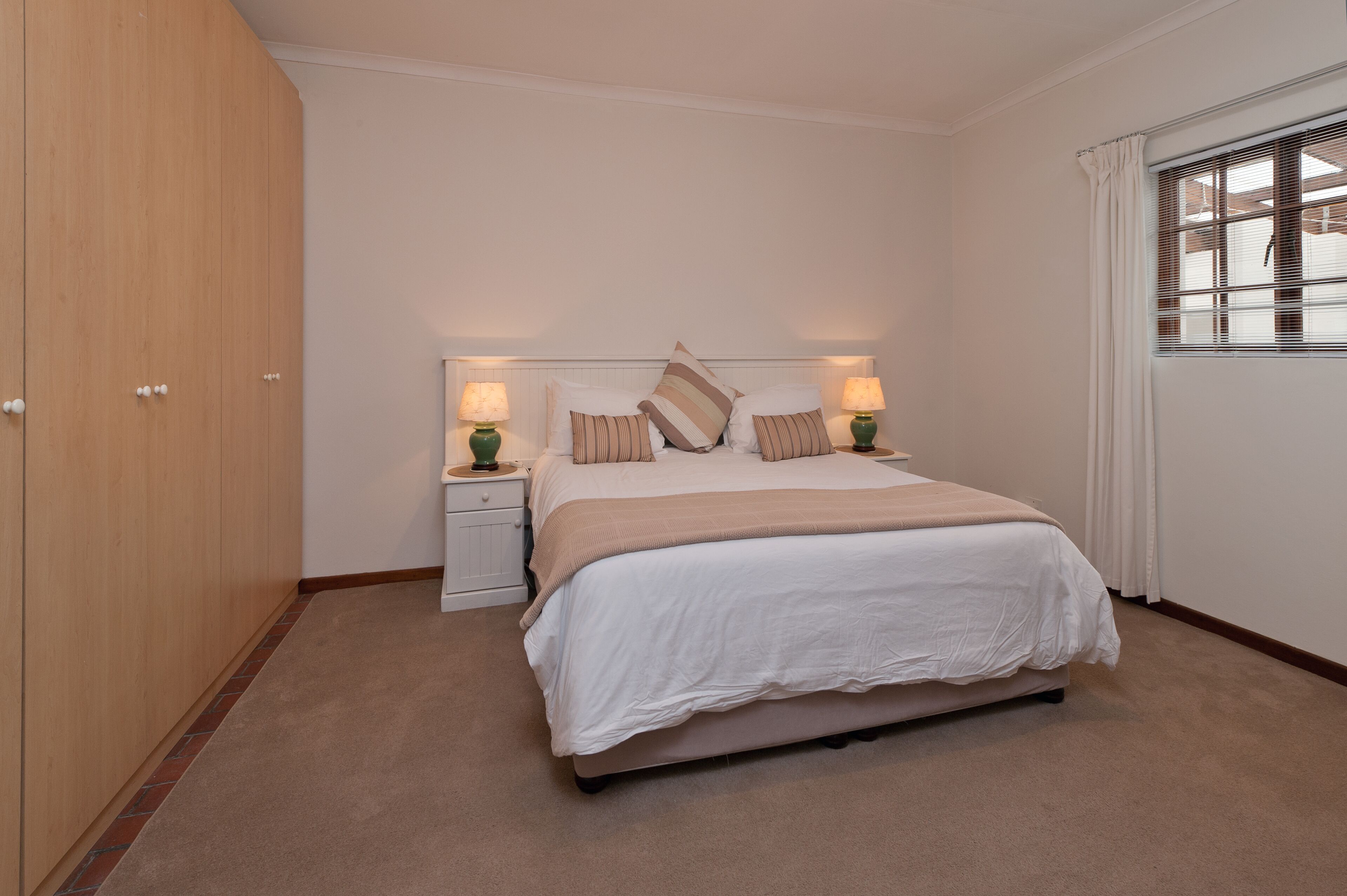 A bedroom with a large bed, two bedside tables with lamps, a headboard, and a window with blinds.