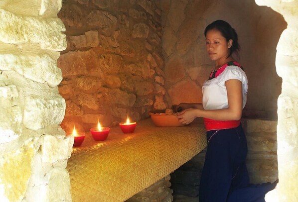 A woman in traditional attire standing in a stone-walled room with a woven bench and lit candles.