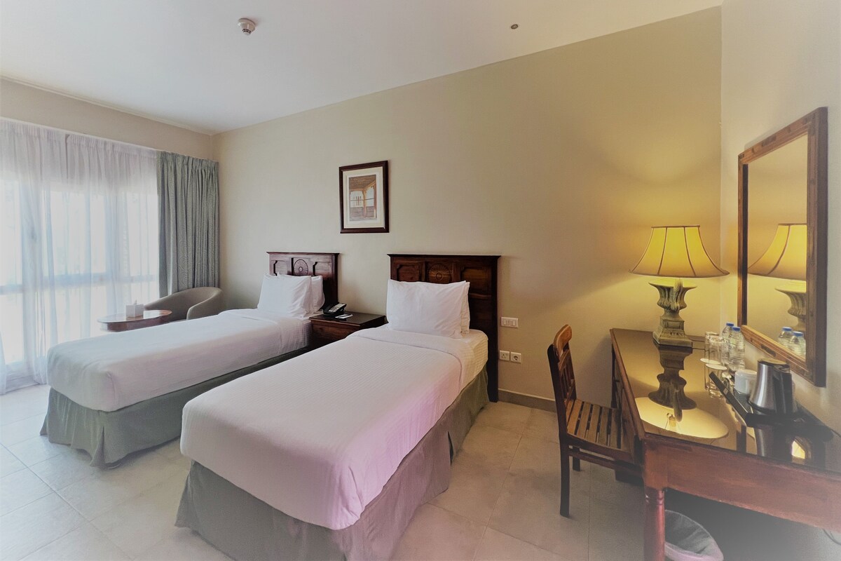 Een hotelkamer met twee bedden, een bureau met lamp, een stoel en een raam met gordijnen.