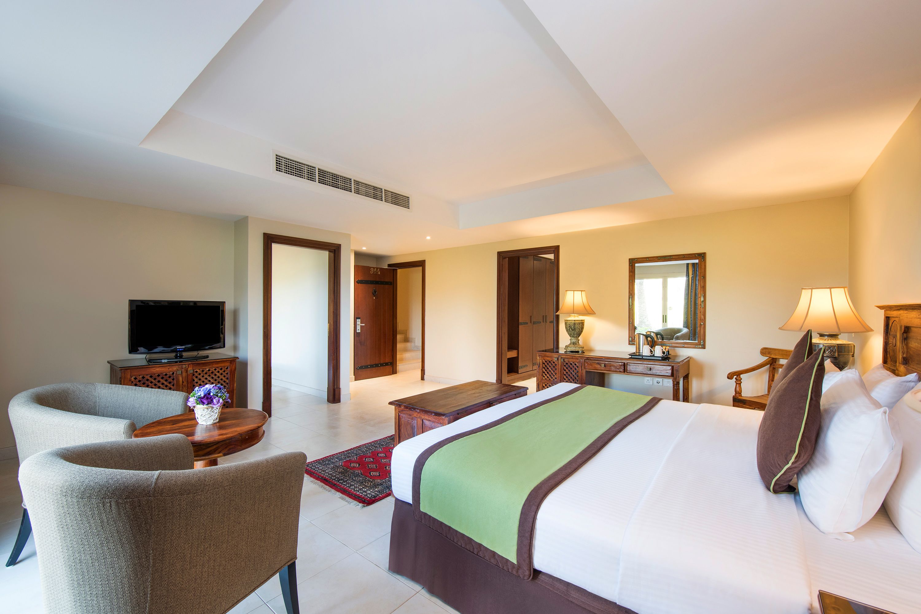 A hotel room with a bed, two armchairs, a television, a desk with a lamp, and a mirror.