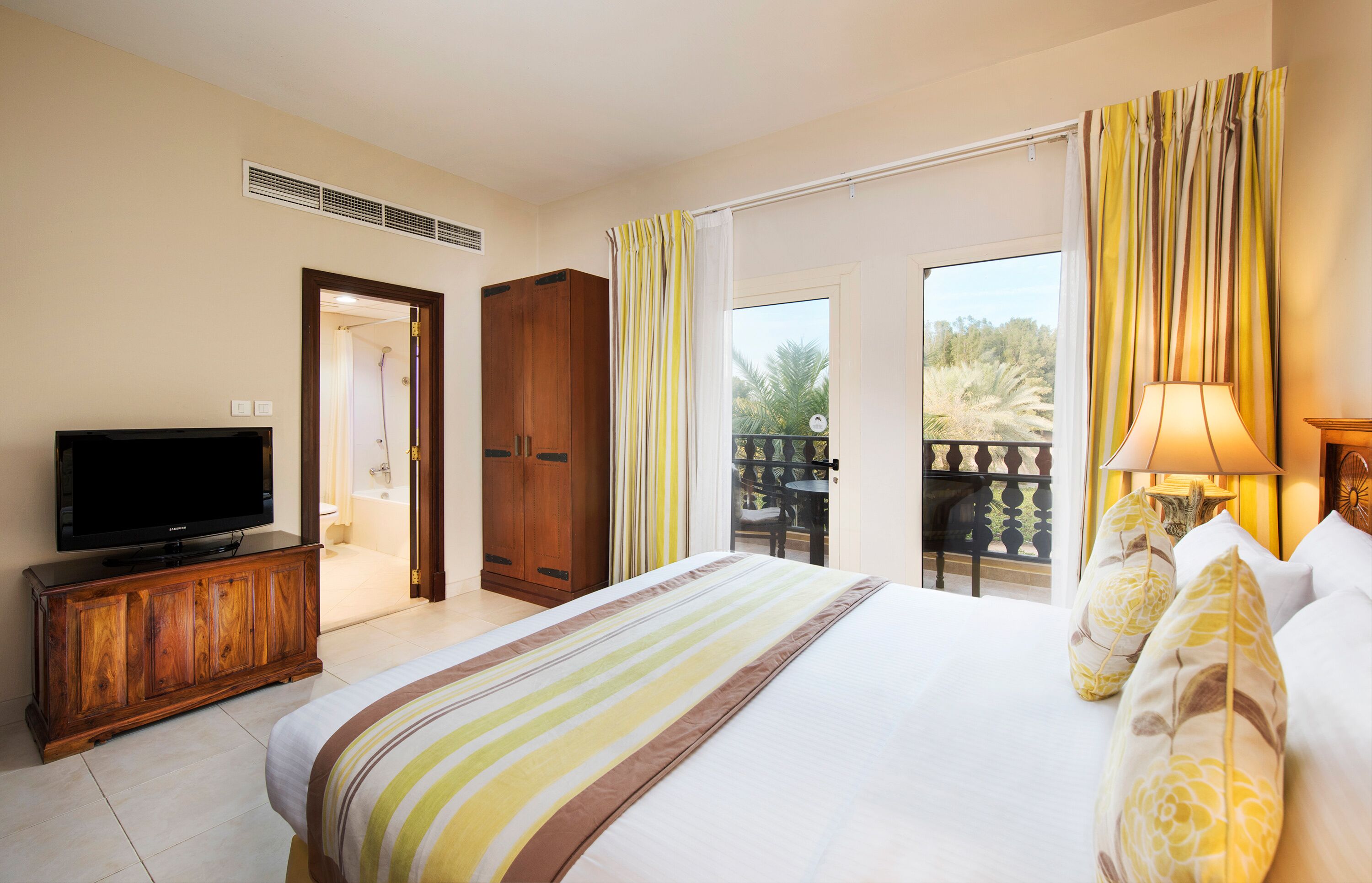 Una habitación de hotel con una cama, un televisor sobre un mueble de madera, un balcón con cortinas amarillas y vistas a palmeras.