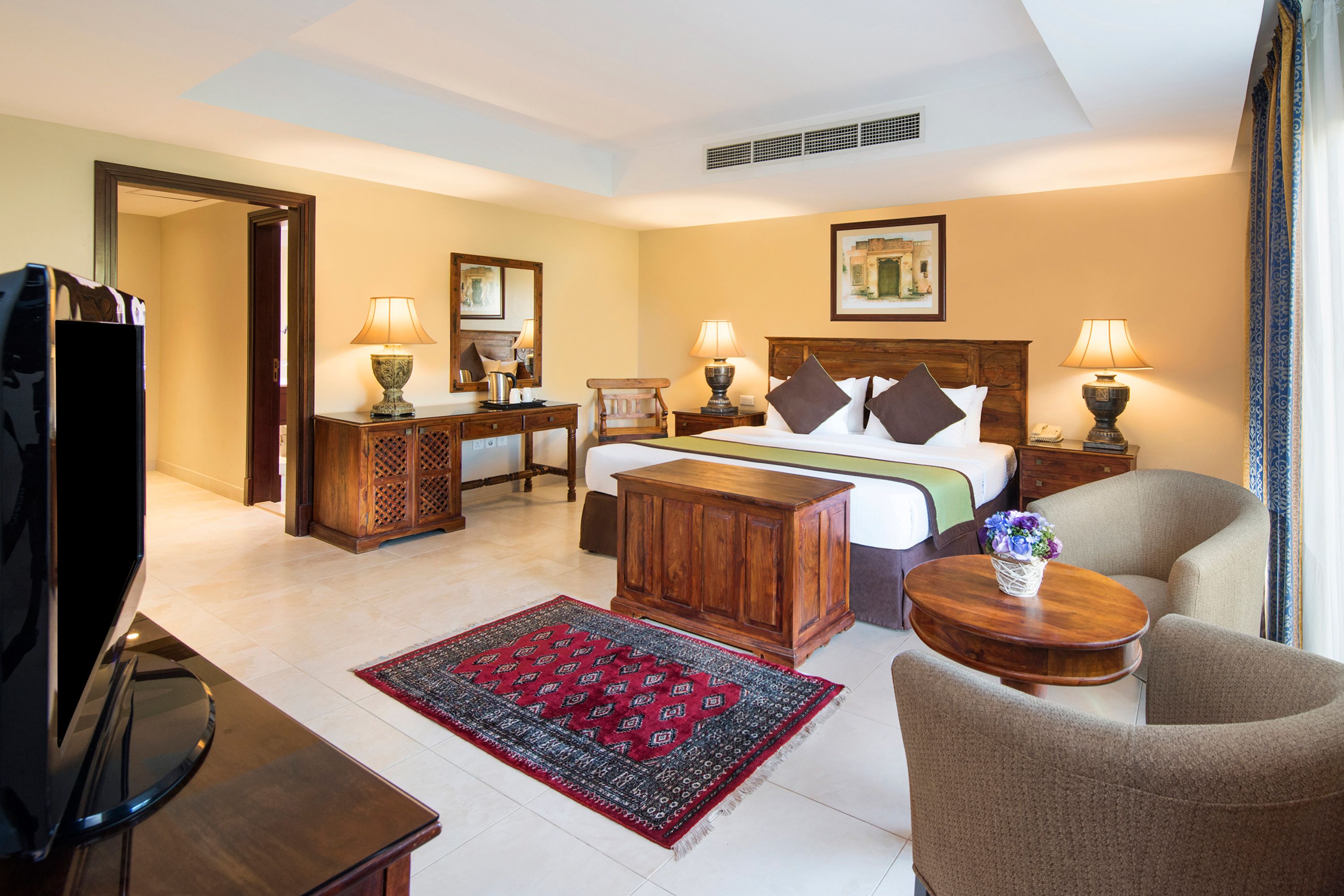 Une chambre d’hôtel comprenant un lit, deux tables de chevet avec des lampes, une télévision, une armoire en bois, un tapis rouge à motifs et une table ronde en bois sur laquelle est posé un vase de fleurs.