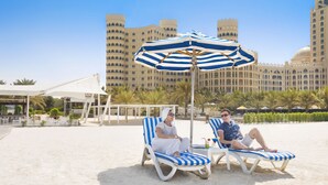 Two lounge chairs with striped cushions on a sandy beach, a large striped umbrella, and a grand hotel with multiple towers in the background. - Al Hamra Village Hotel (Ras Al Khaimah)