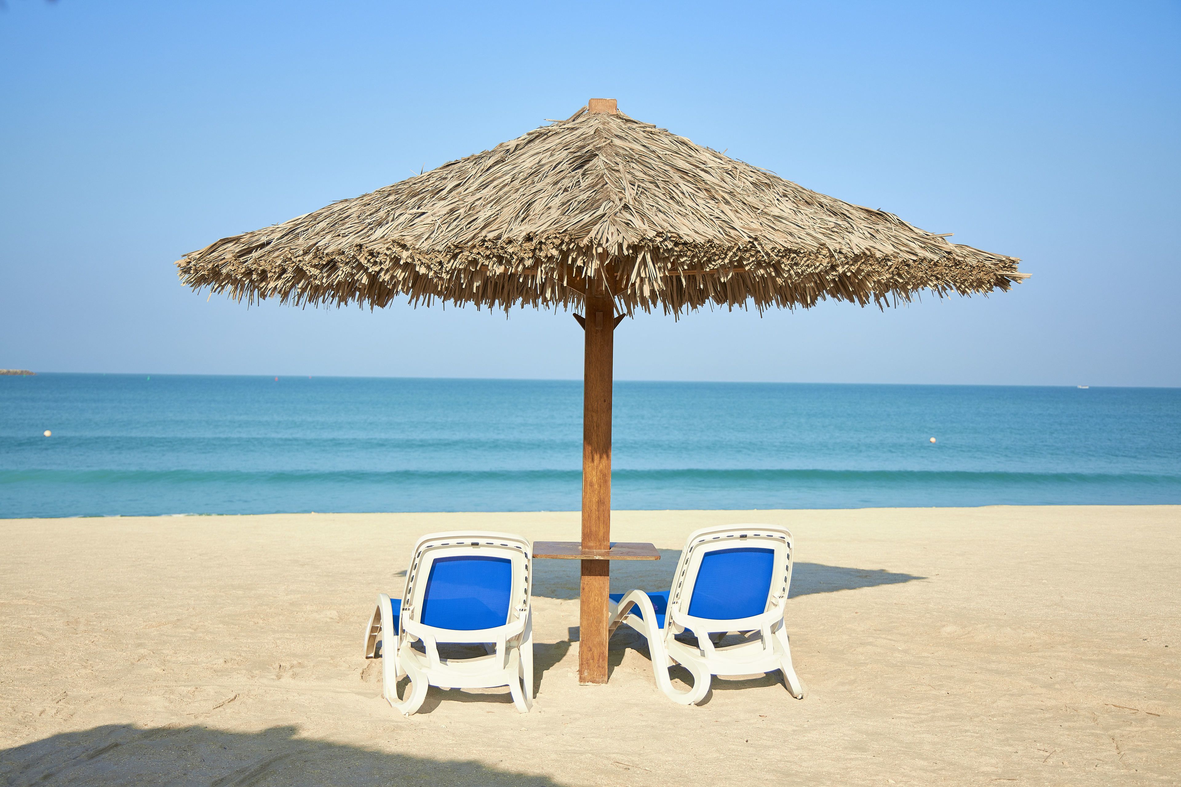 A beach with two white lounge chairs under a thatched umbrella, a wooden table, and the ocean in the background.