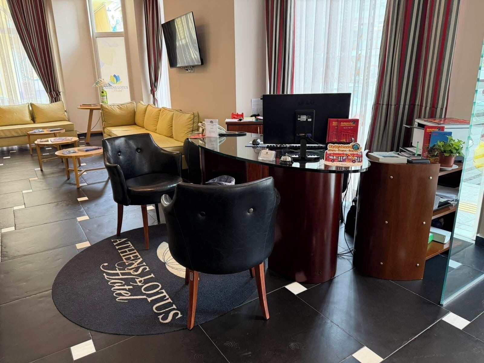 A hotel lobby with a sofa, two black chairs, a glass table, a TV, and a sign for 'Athens Lotus.'