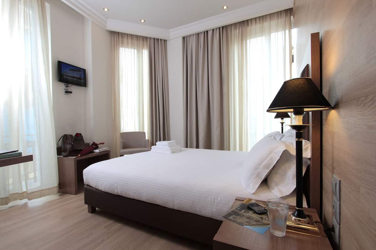 A hotel room with a bed, a desk, a chair, a lamp, and a TV on the wall.