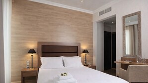 A hotel room with a large bed, two bedside lamps, a wooden headboard, a desk with a chair, a mirror, and a small table with a pitcher and glasses. - Athens Lotus Hotel (Athens)