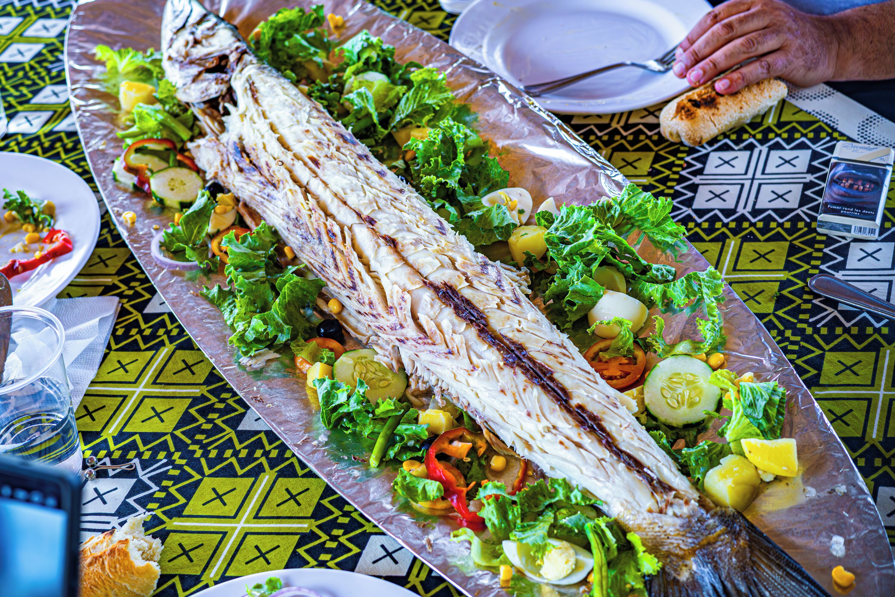 A large grilled fish surrounded by vegetables on a patterned tablecloth.