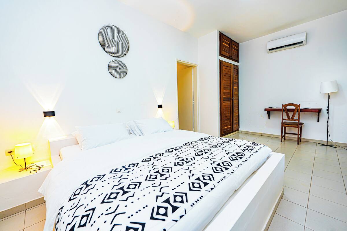 A hotel room with a bed, bedside lamps, a desk, and an air conditioning unit.