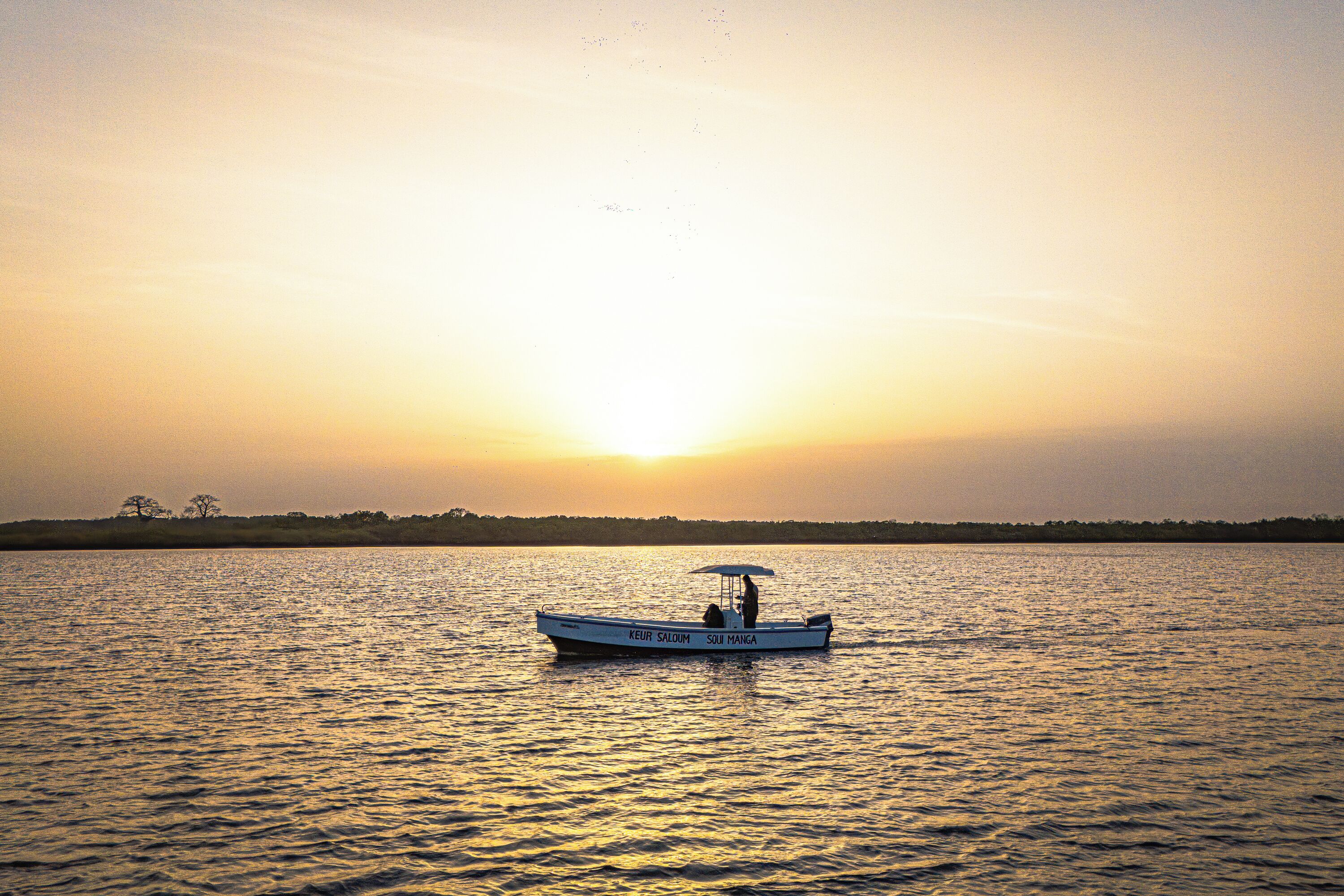 A boat with the text "KEUR SALOM" and "SUI MANGA" on its side, floating on calm water during sunset.