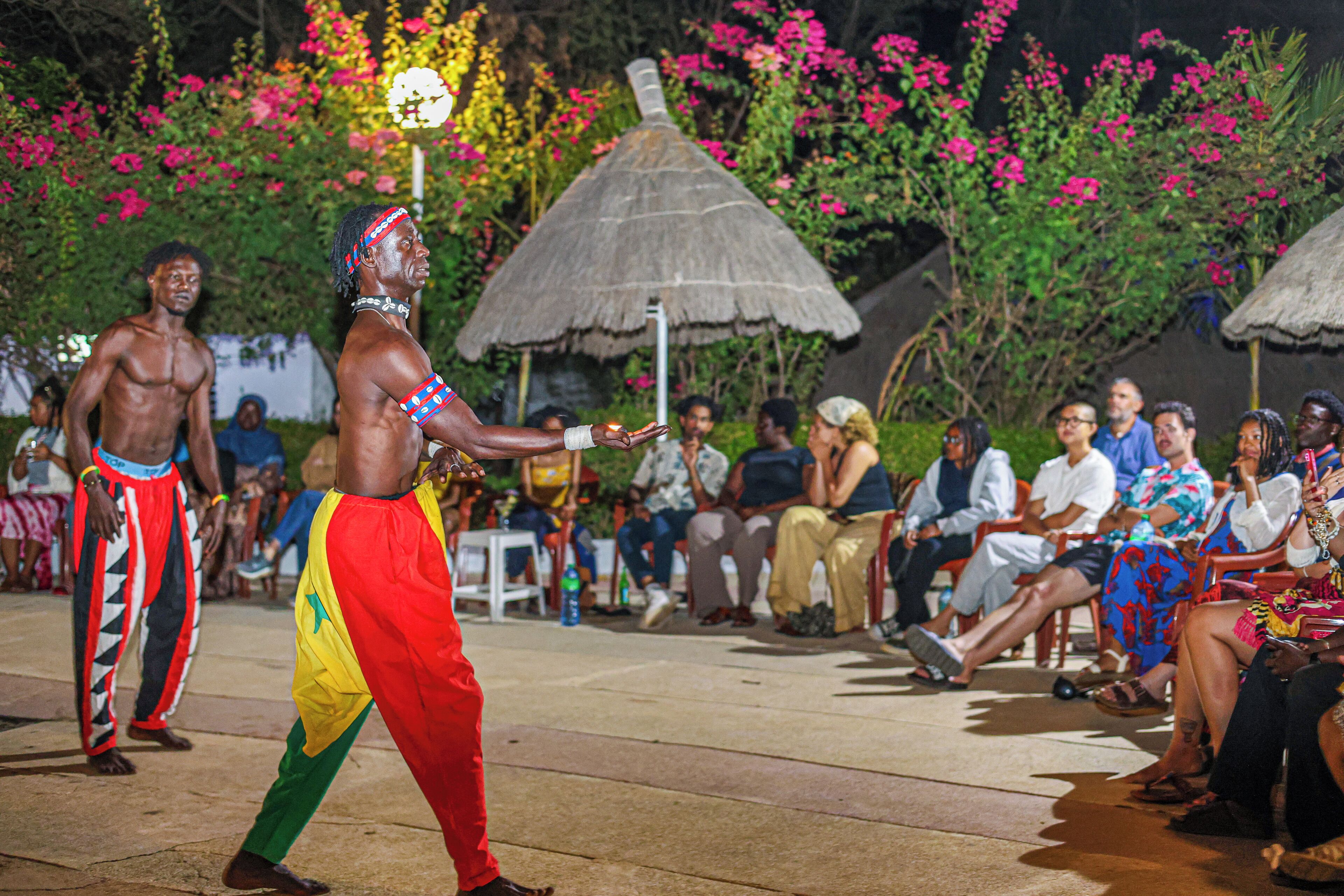 A performer in traditional attire with a thatched-roof hut and pink flowers in the background.