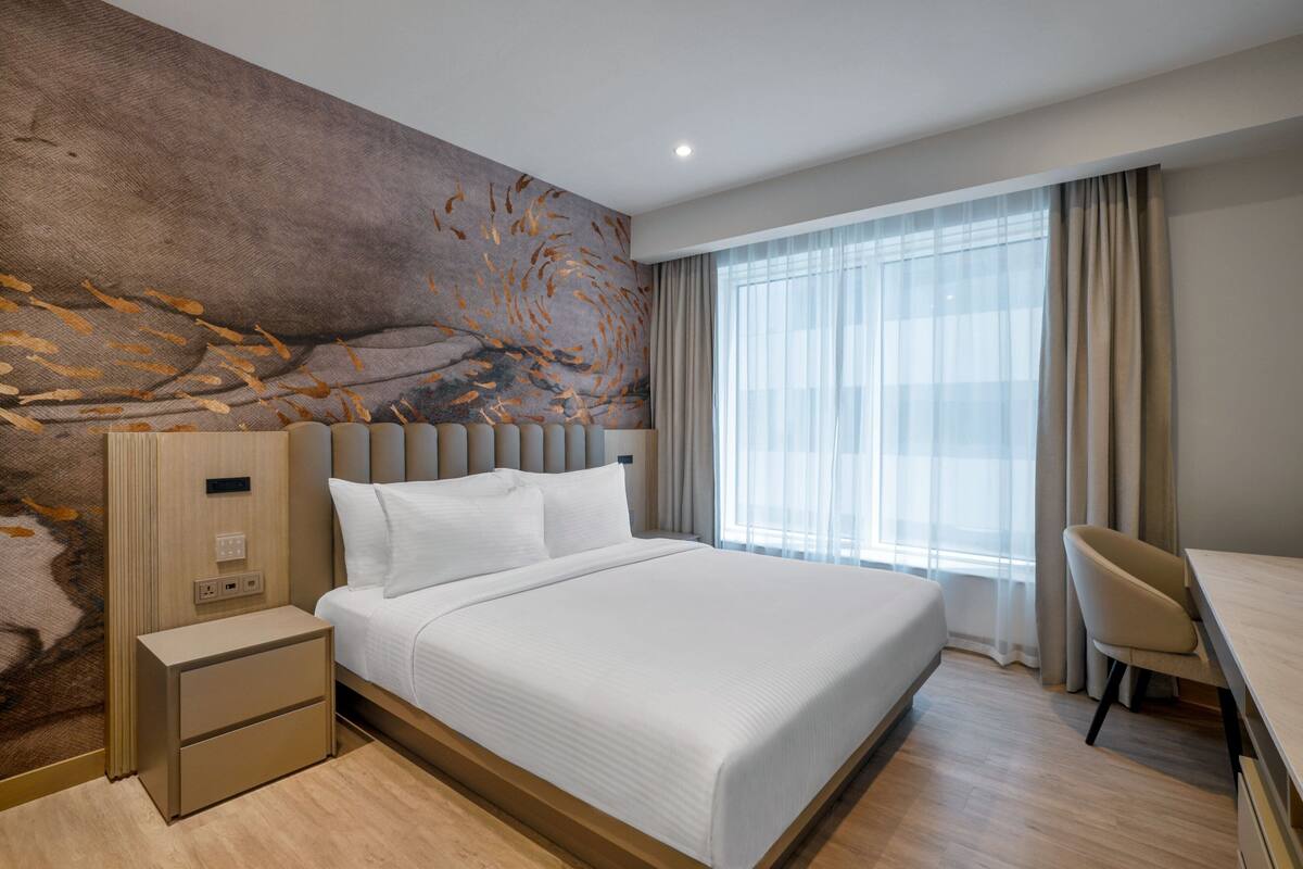 A modern hotel room with a large bed, a desk, and a view of the city through large windows.
