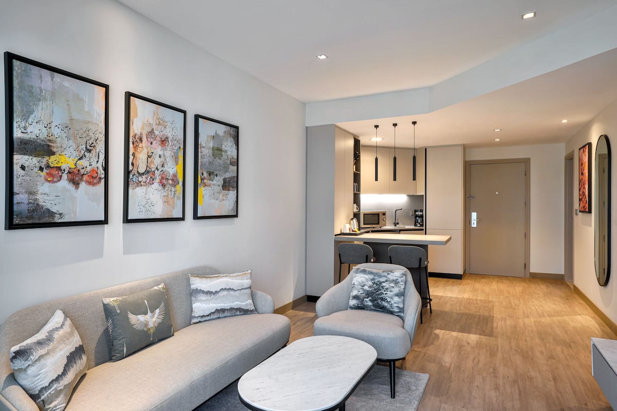 A modern living room with a sofa, armchair, and coffee table. There is a kitchen area with a microwave and cabinets.