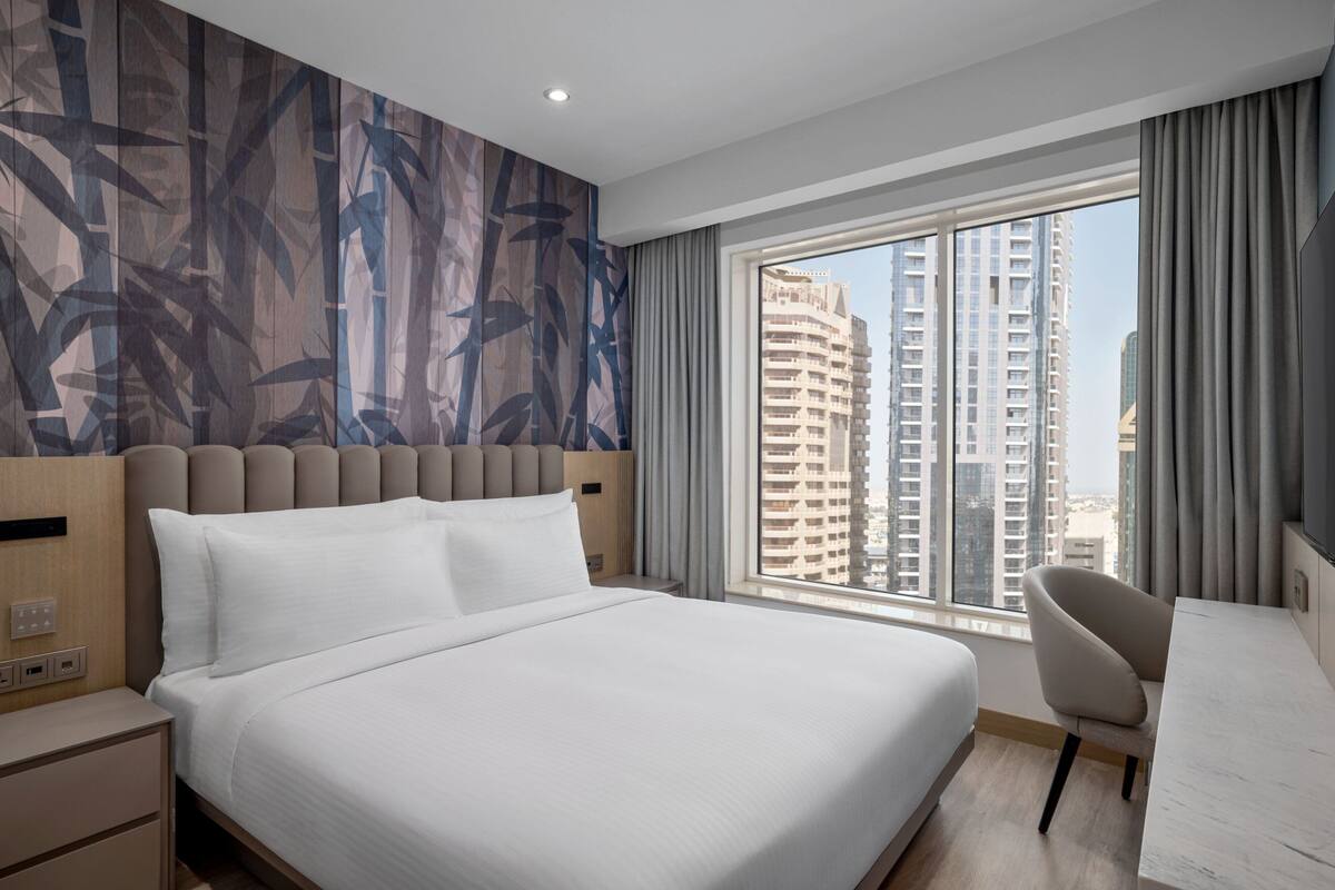 A modern hotel room with a large bed, a desk, and a view of tall buildings.