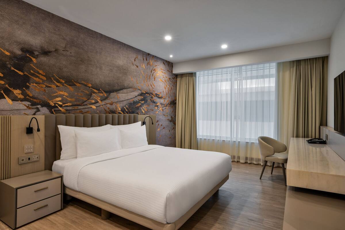 A modern hotel room with a large bed, a desk, and a view of the city through the window.
