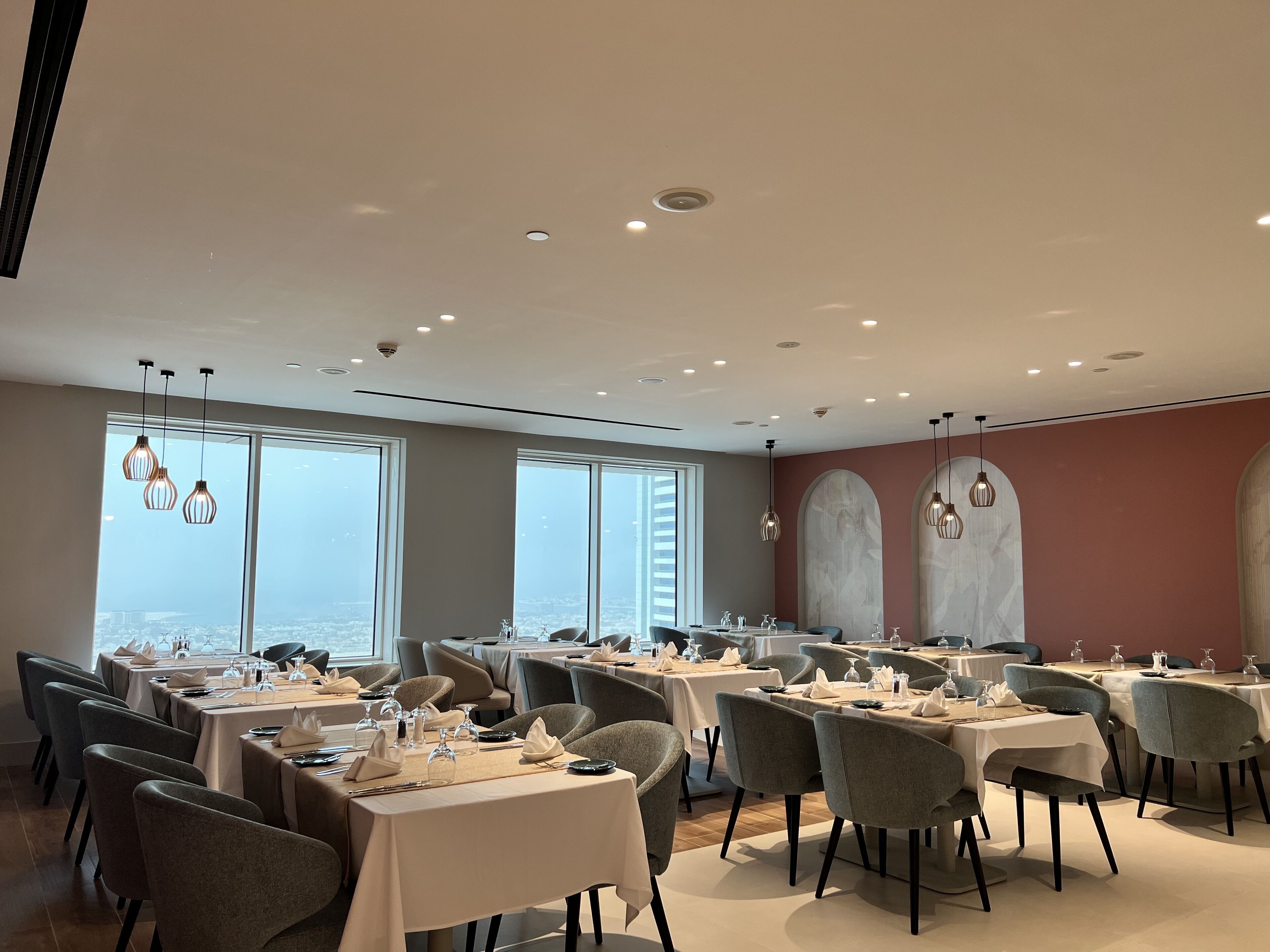 A modern dining area with tables set for service, large windows offering a city view, and contemporary lighting fixtures.