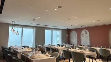 A modern dining area with tables set for service, large windows offering a city view, and contemporary lighting fixtures.